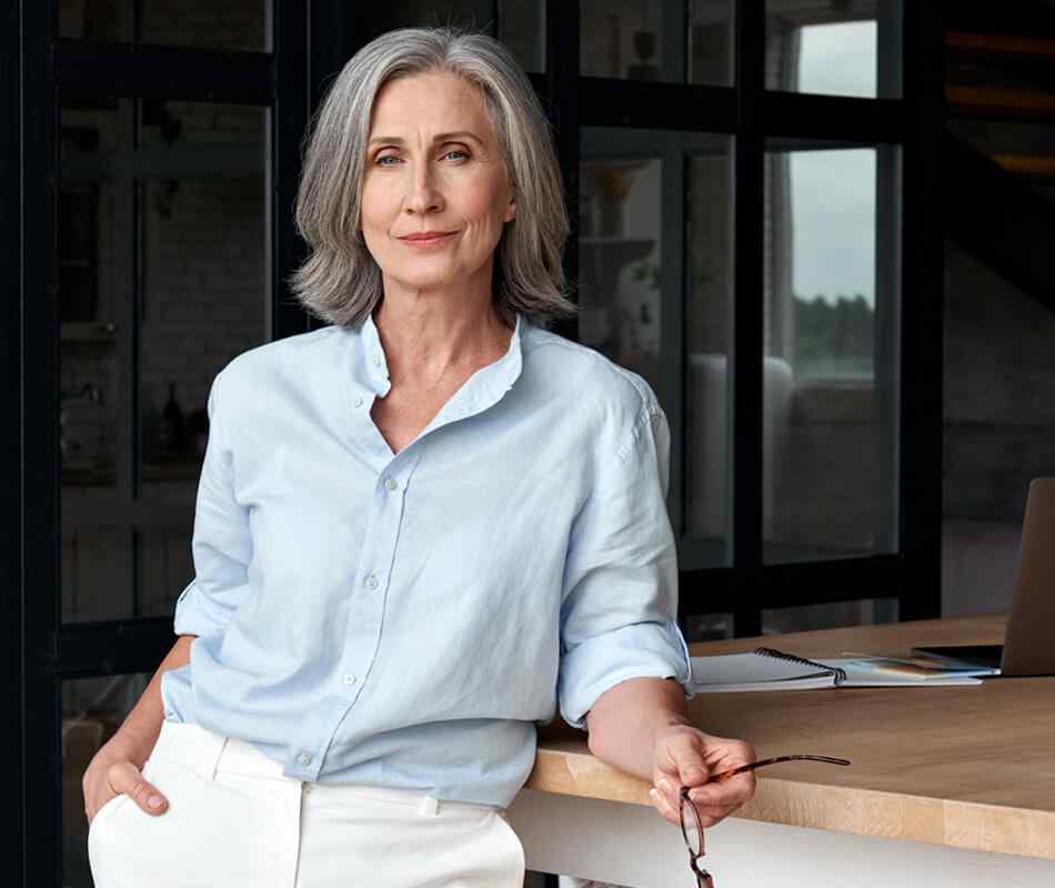 Confident stylish european mature middle aged woman standing at workplace. Stylish older senior businesswoman, 60s gray-haired lady executive leader manager looking at camera in office, portrait.